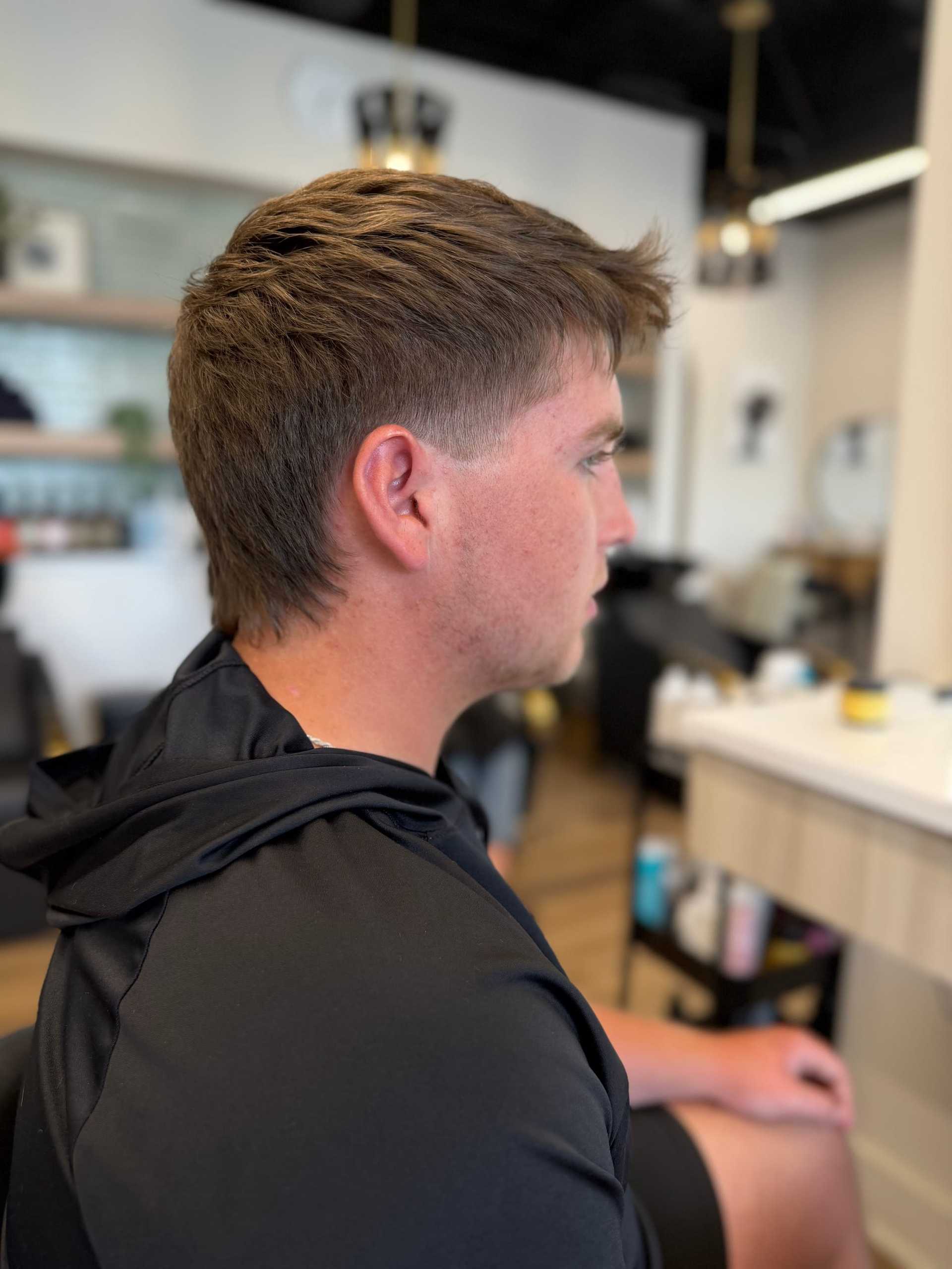 Man in a black shirt seated in a hair salon, showing a side profile with a fresh haircut.