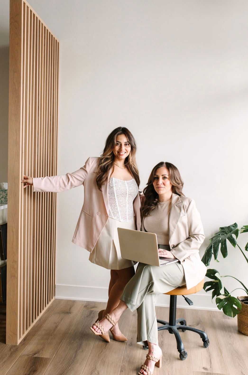 Two women in stylish outfits, one seated with a laptop, posing in a modern, minimalist room.