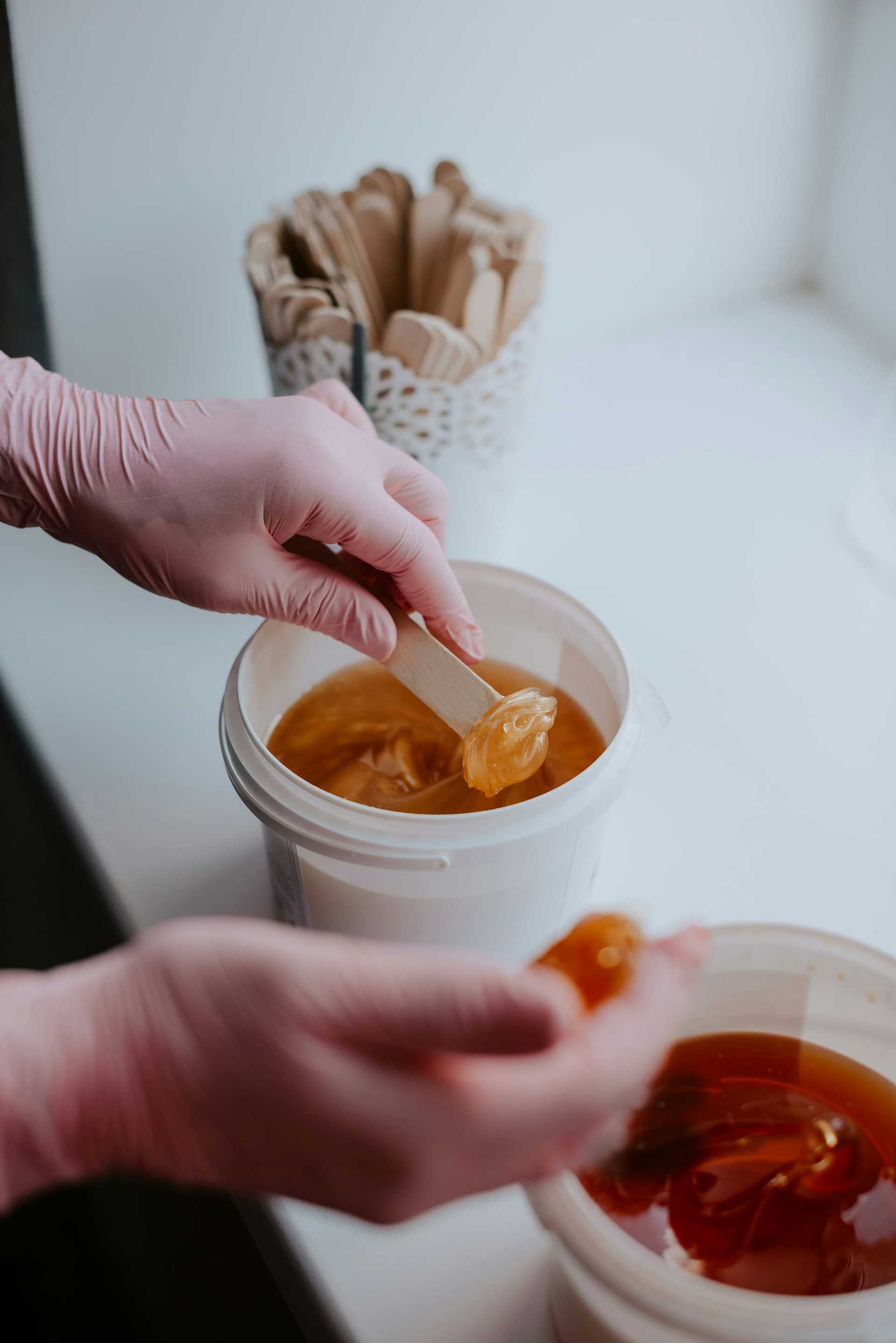 Hands wearing pink gloves apply wax with a wooden spatula from a bucket, suggesting hair removal.